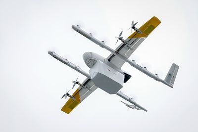 Witte bezorgdrone met gele vleugels in vlucht tegen grijze lucht.