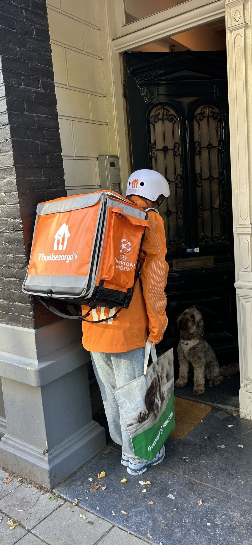 Thuisbezorgd.nl bezorger met oranje rugtas en Maxi Zoo tas aan voordeur met hond.