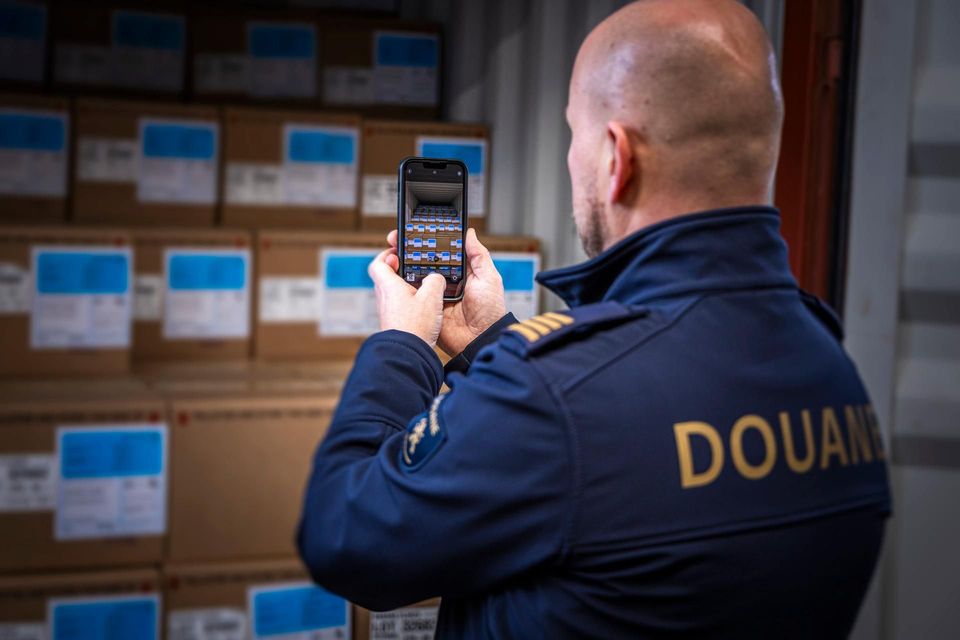 Douanier fotografeert stapels kartonnen dozen in container met smartphone.