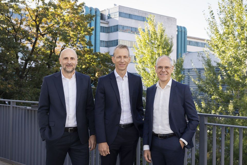 Drie mannelijke bestuurders van TGW Logistics poseren buiten voor een industrieel gebouw.