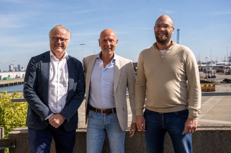 Drie mannen poseren buiten met een haven op de achtergrond.