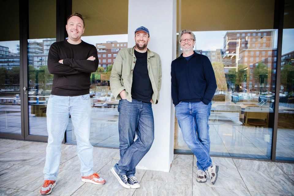 Drie mannen poseren lachend buiten een kantoorgebouw aan het water.