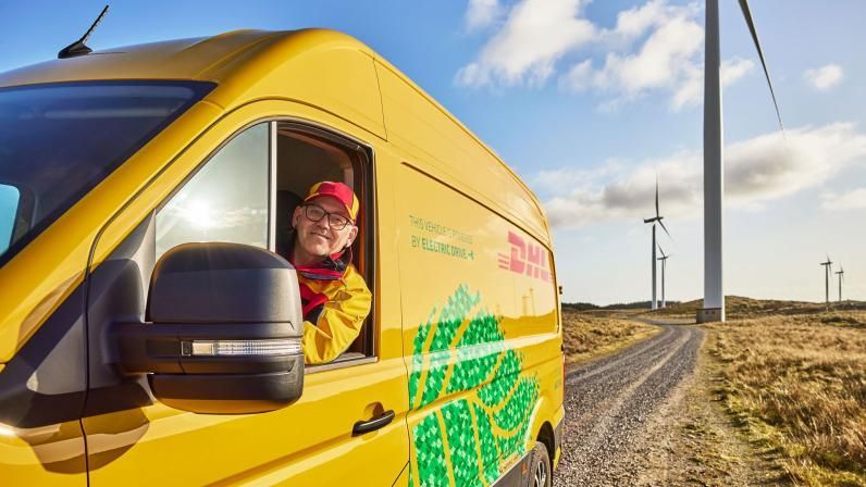 DHL-chauffeur leunt uit raam van gele elektrische bestelbus bij windmolens.