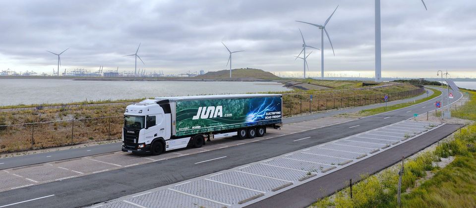 Elektrische JUNA-vrachtwagen op kustweg met windmolens en haven op achtergrond.