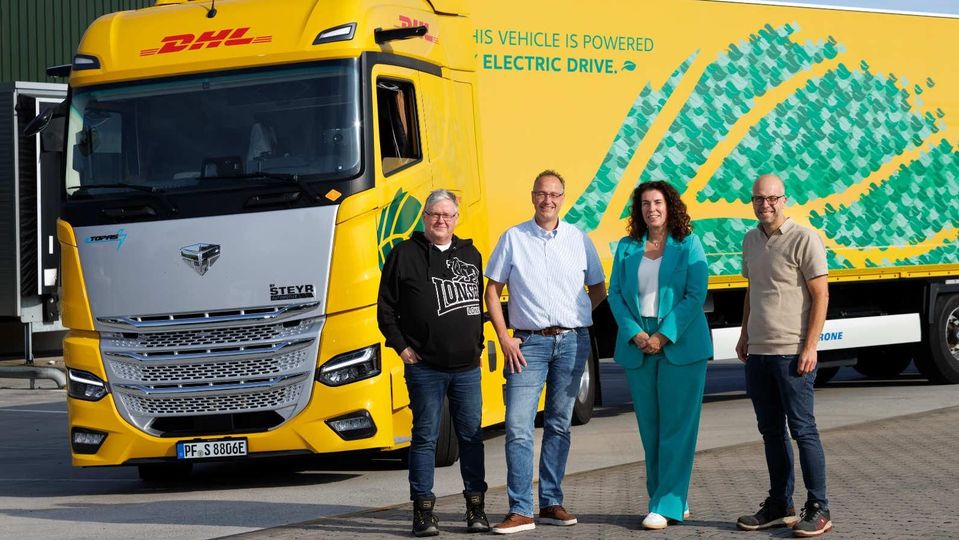 Vier mensen poseren voor gele elektrische DHL-vrachtwagen op parkeerterrein.