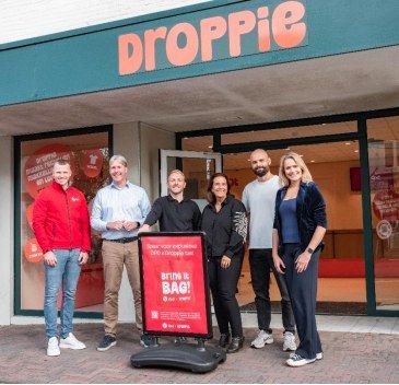Zes personen staan voor een Droppie-winkel met campagnebord.
