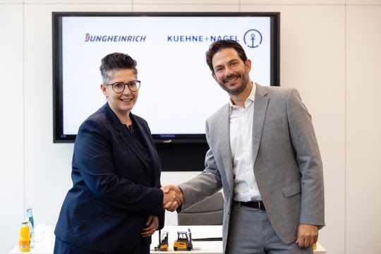Man en vrouw schudden handen voor scherm met logo's Jungheinrich en Kuehne+Nagel.