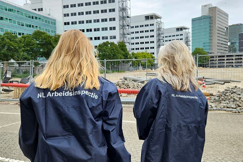 Twee inspecteurs met NL Arbeidsinspectie-jas op de rug, bouwplaats op achtergrond.