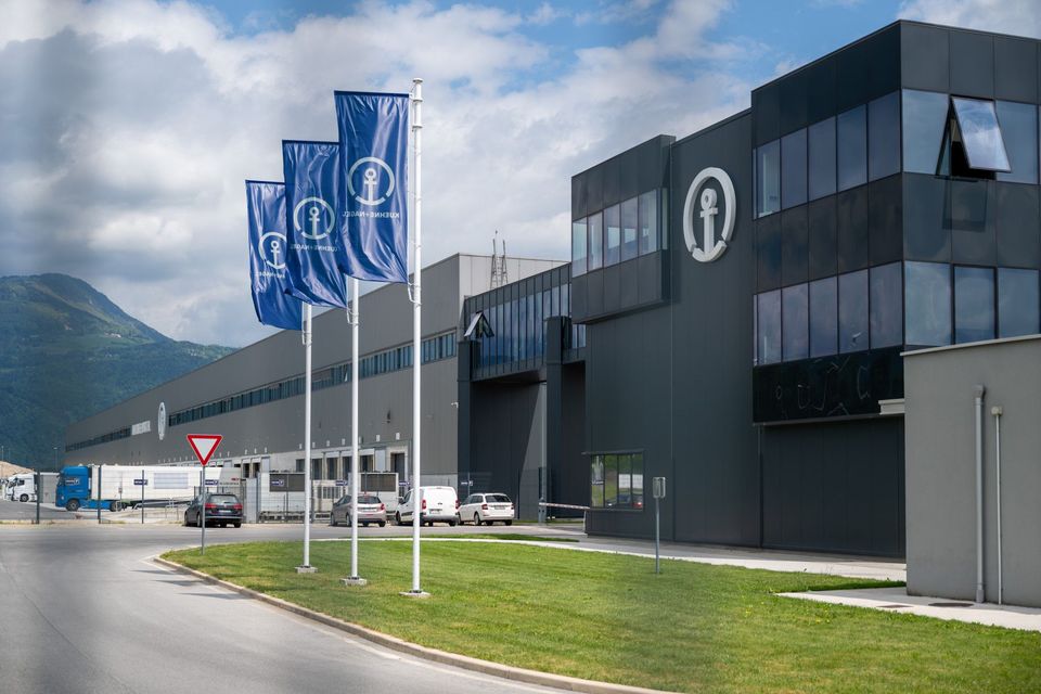 Exterieur van Kuehne+Nagel logistiek centrum met blauwe vlaggen en bergen op achtergrond.