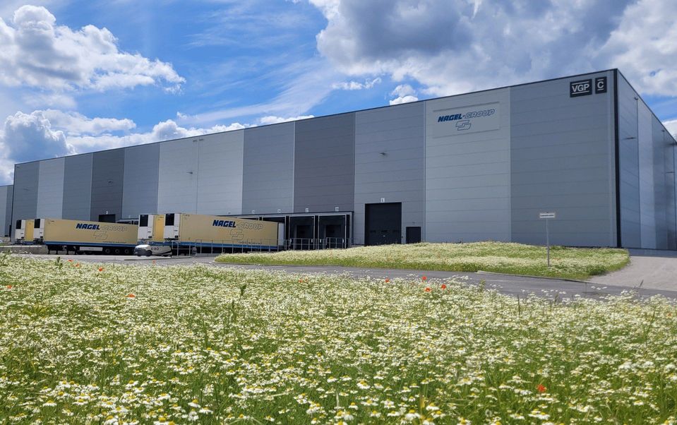 Grijze logistiekhal met Nagel-Group-opleggers aan laaddocks en bloeiende weide.