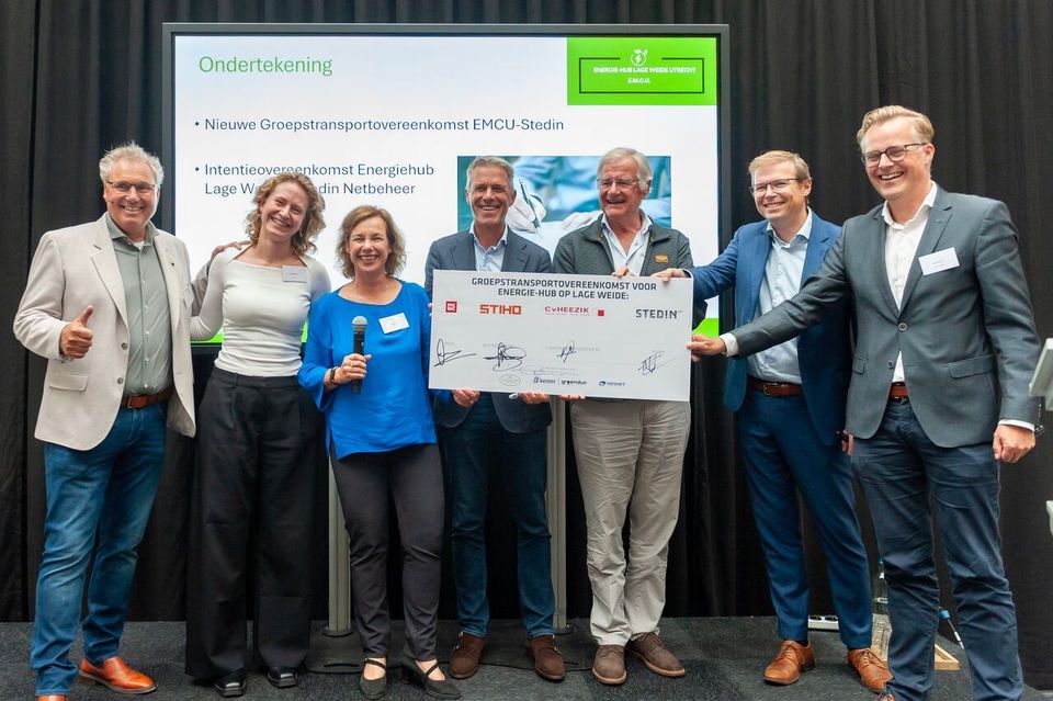 Zeven mensen houden gezamenlijk een ondertekend overeenkomstbord vast op podium.