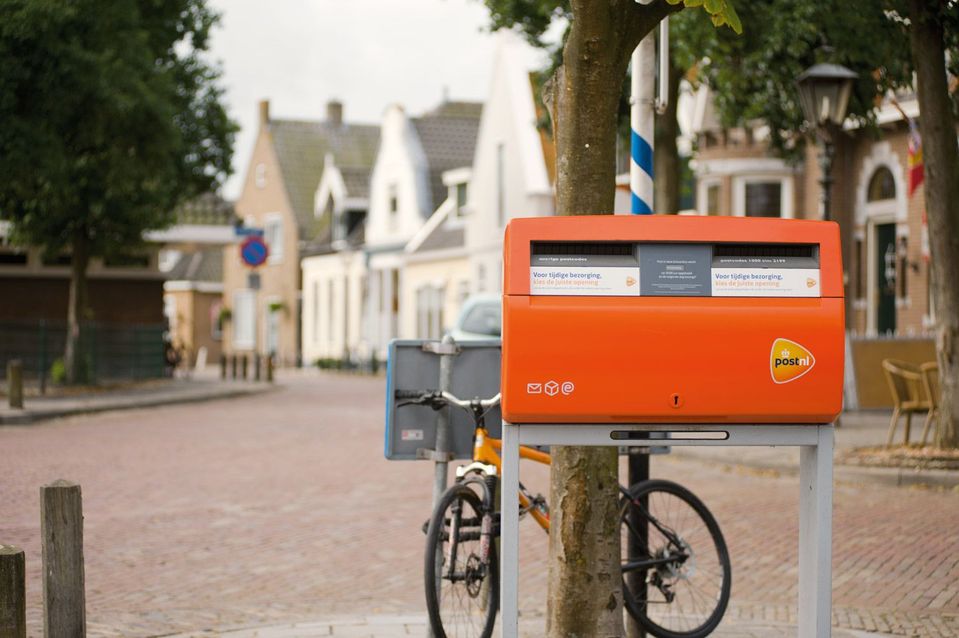 Oranje PostNL-brievenbus op een dorpsplein met fietsen en woningen op de achtergrond.