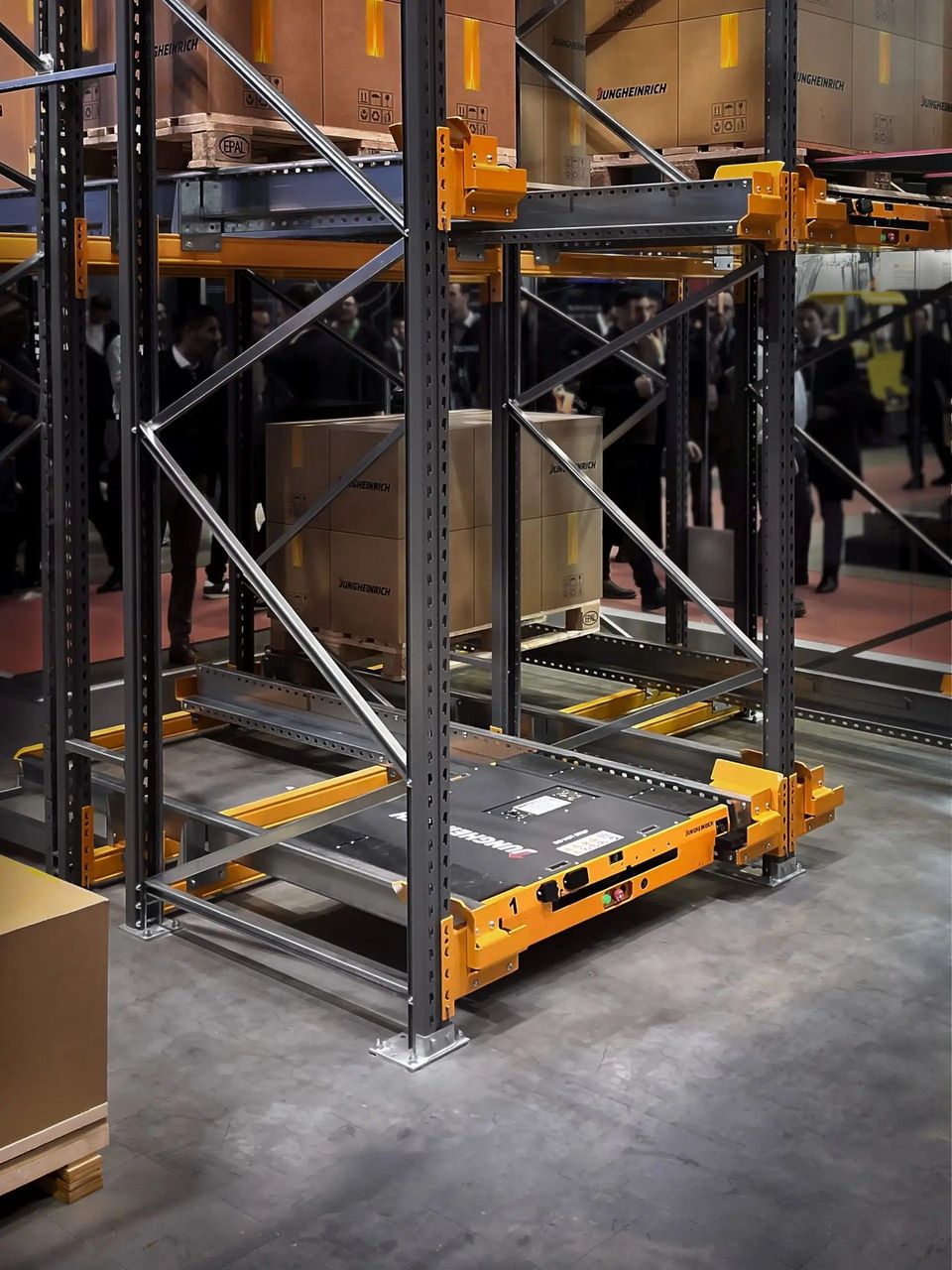Gele palletshuttle in compacte palletstelling met dozen op beurs.