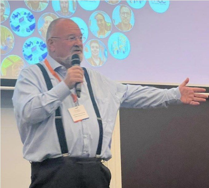 Man met microfoon en bretels spreekt op een podium tijdens conferentie.