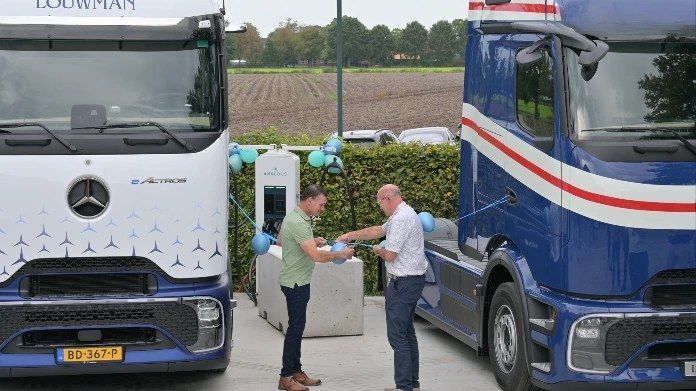 Twee mannen schudden handen bij laadpaal tussen twee elektrische vrachtwagens.
