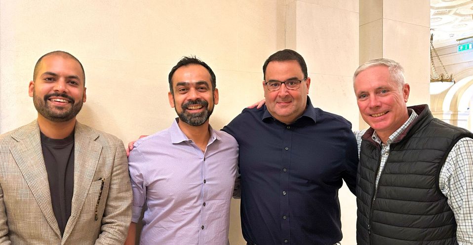 Vier lachende mannen poseren samen voor een lichte muur.