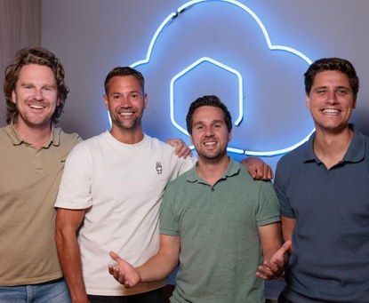 Vier lachende mannen staan voor een blauw neon wolklogo.