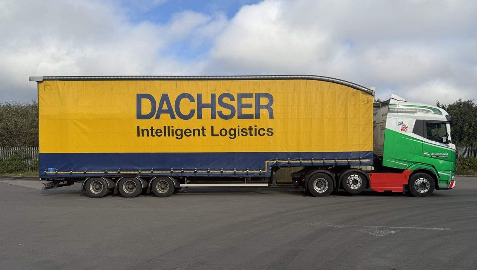Groene Drummond-truck met geel-blauwe DACHSER schuifzeiloplegger op parkeerplaats.
